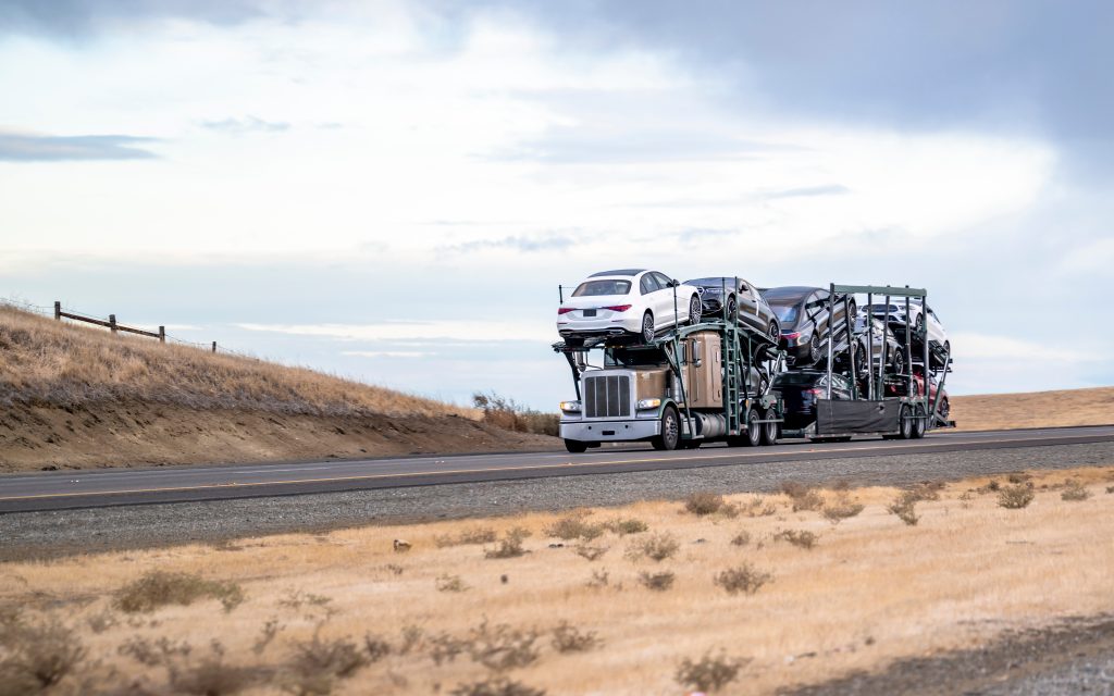 Beige commercial big rig car hauler semi truck with turned on headlight transporting fastened cars on two level hydraulic modular semi trailer running on the highway road in Idaho at twilight