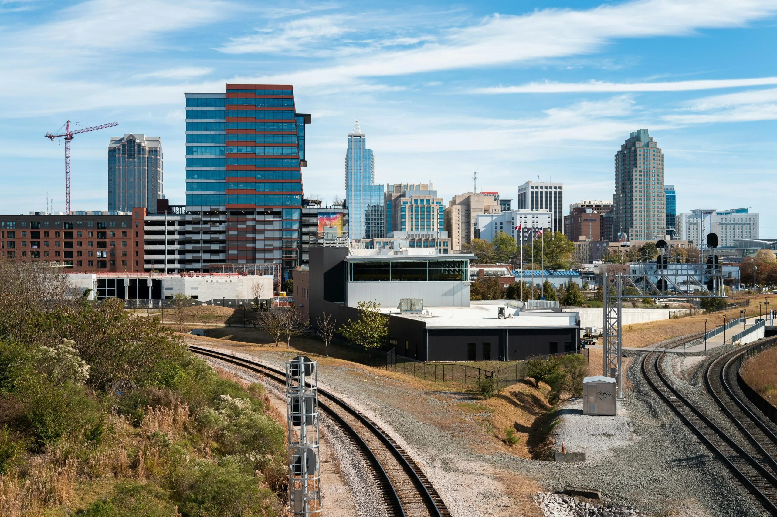 A stunning aerial view of the Raleigh skyline, showcasing modern skyscrapers, surrounding greenery, and a blue sky with wispy clouds.
